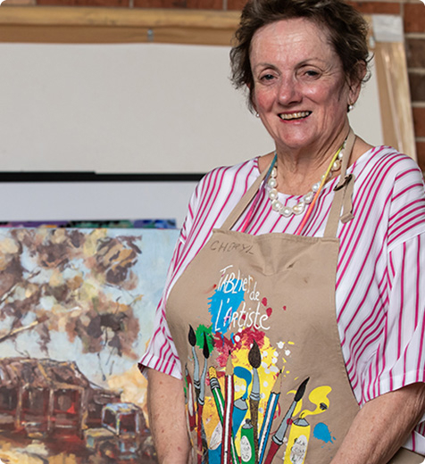 Cheryl is smiling to camera, standing in front of her artwork, and wearing an art apron