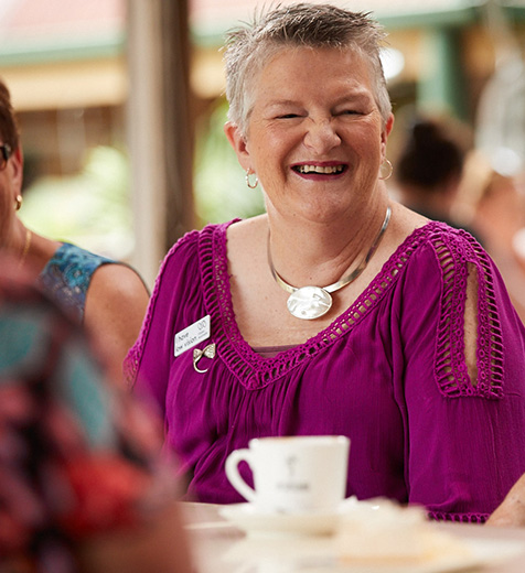 Norma sits at a table with a cup of tea, smiling and socialising with others.