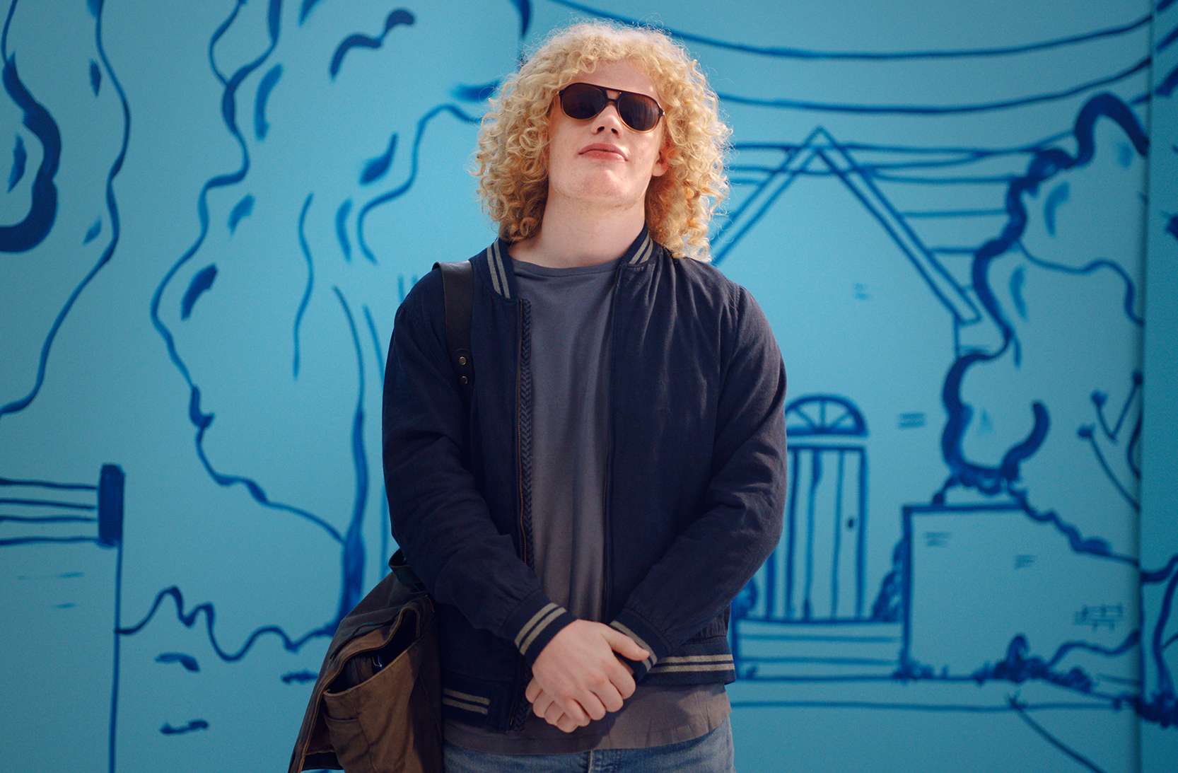 A young man with curly blonde hair dressed in jeans and a bomber jacket stands proudly with his hands folded in front of him, wearing sunglasses. He is standing in front of a painted light blue and navy outdoor scene.