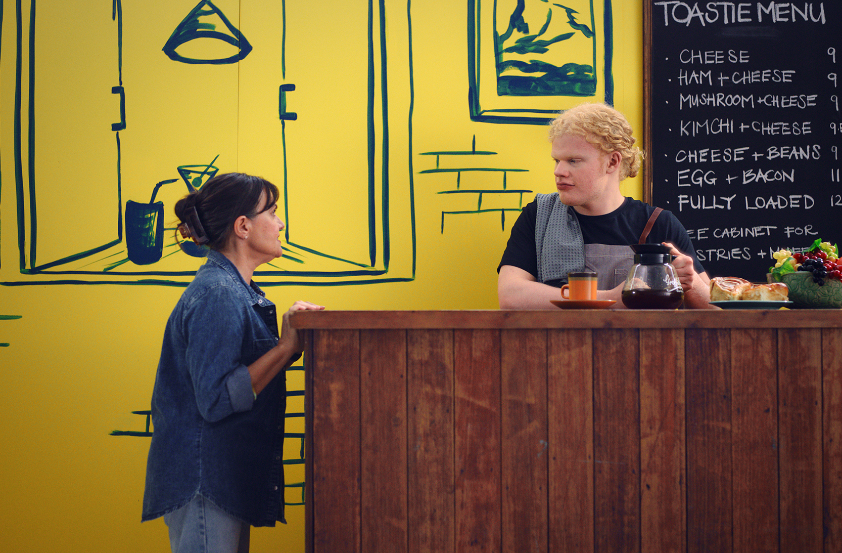 A woman wearing a denim shirt and jeans is standing at a wooden bar, speaking to a blonde man in a black t-shirt and grey apron behind the bar. There is a blackboard menu behind him with a list of 7 cheese toastie options. They are both in front of a yellow and navy painted café scene.